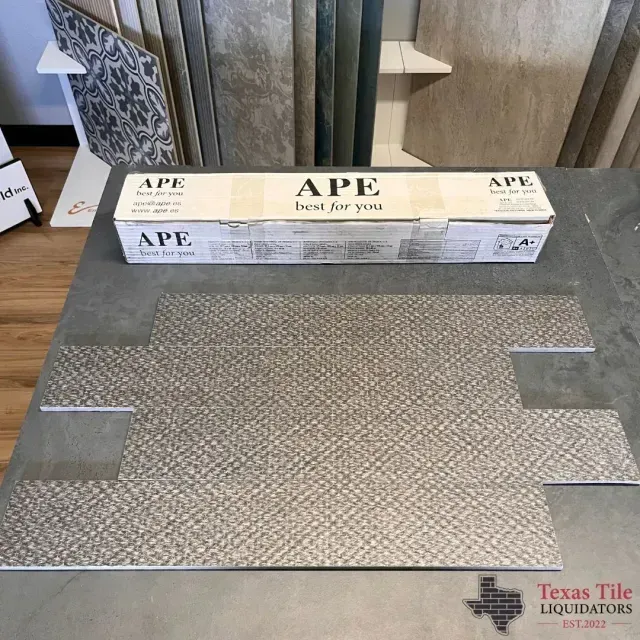 A gray, textured rectangular tile sits on a table in front of an APE-branded box at a tile showroom.