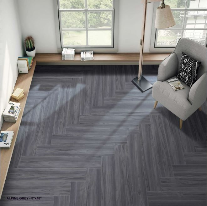 A gray herringbone-patterned floor in a bright, modern room with a window, a wooden ledge with books, and a lounge chair.