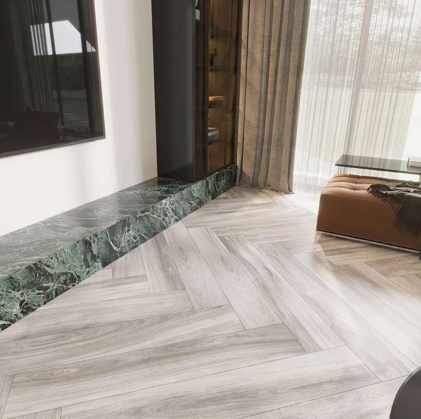 A room with light-colored herringbone wood flooring, a green marble hearth, and a brown ottoman near sheer curtains.