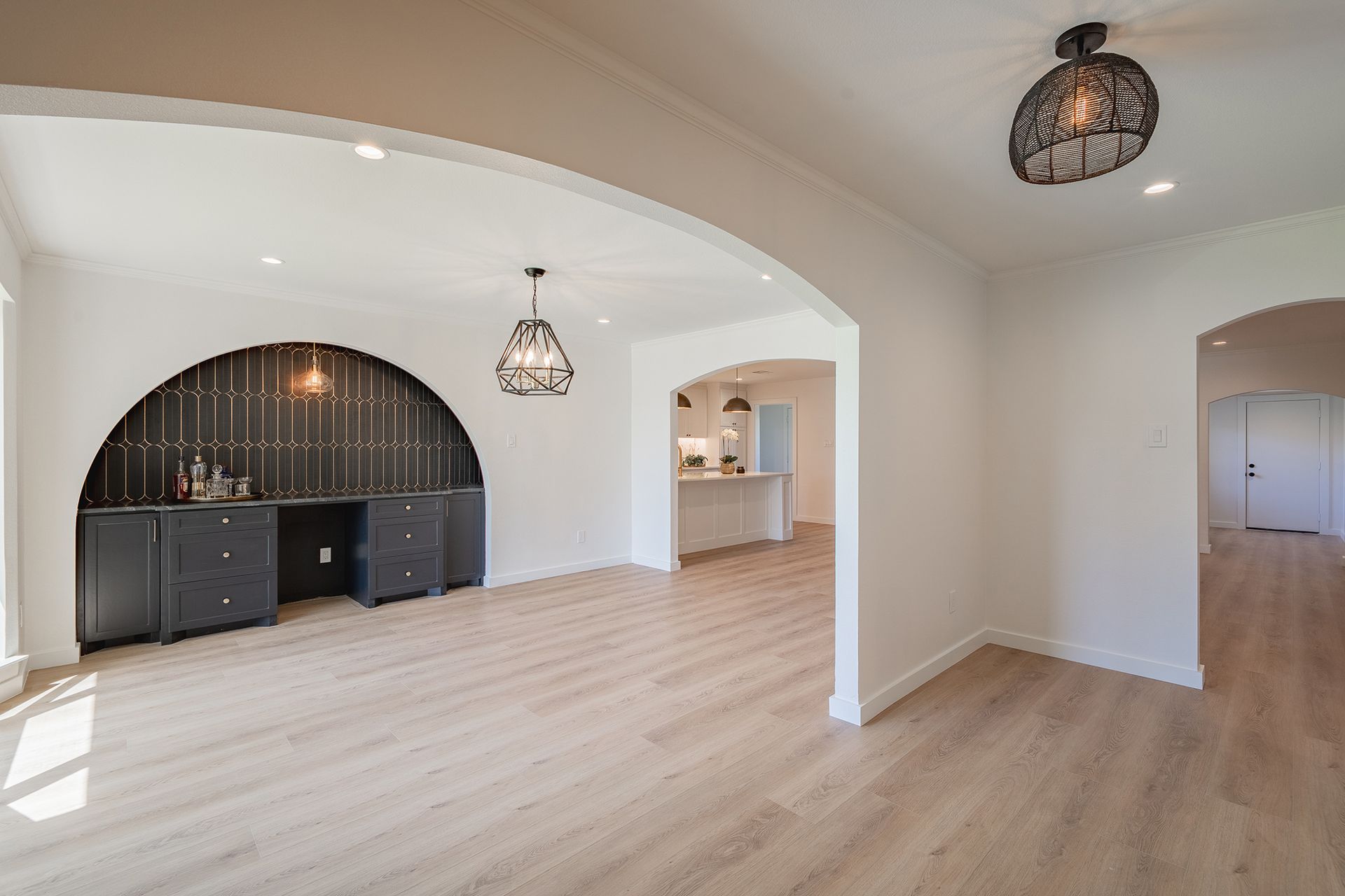 A light-filled room with light wood floors, arched architectural details, a dark built-in desk, and modern pendant lights.