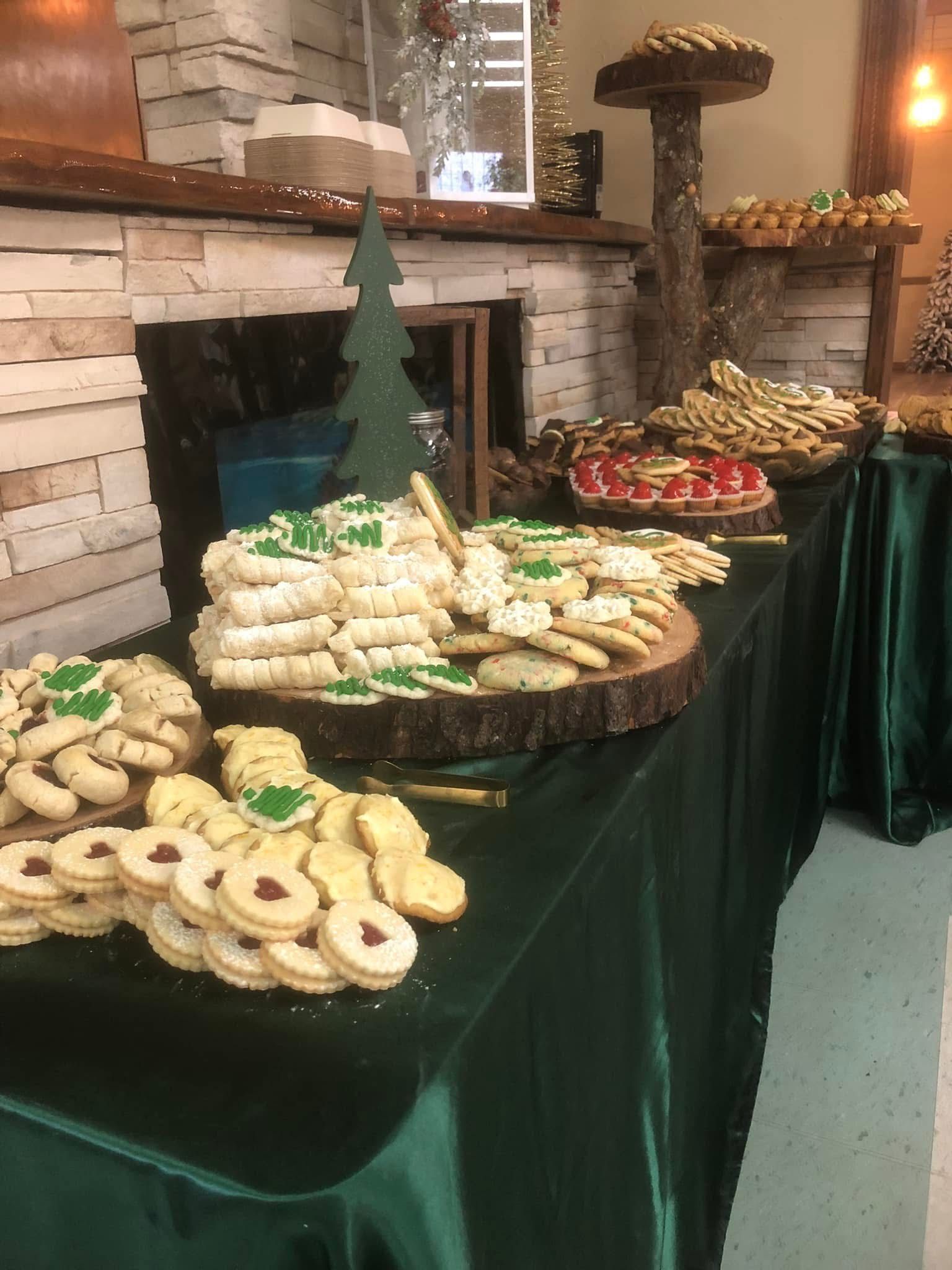 A table laden with holiday cookies on a forest green tablecloth, decorated with holly leaves and Christmas trees.