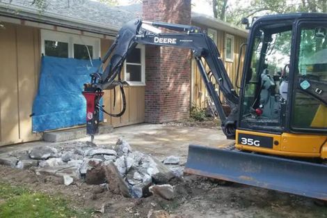 A small excavator is breaking up a concrete patio in front of a house.