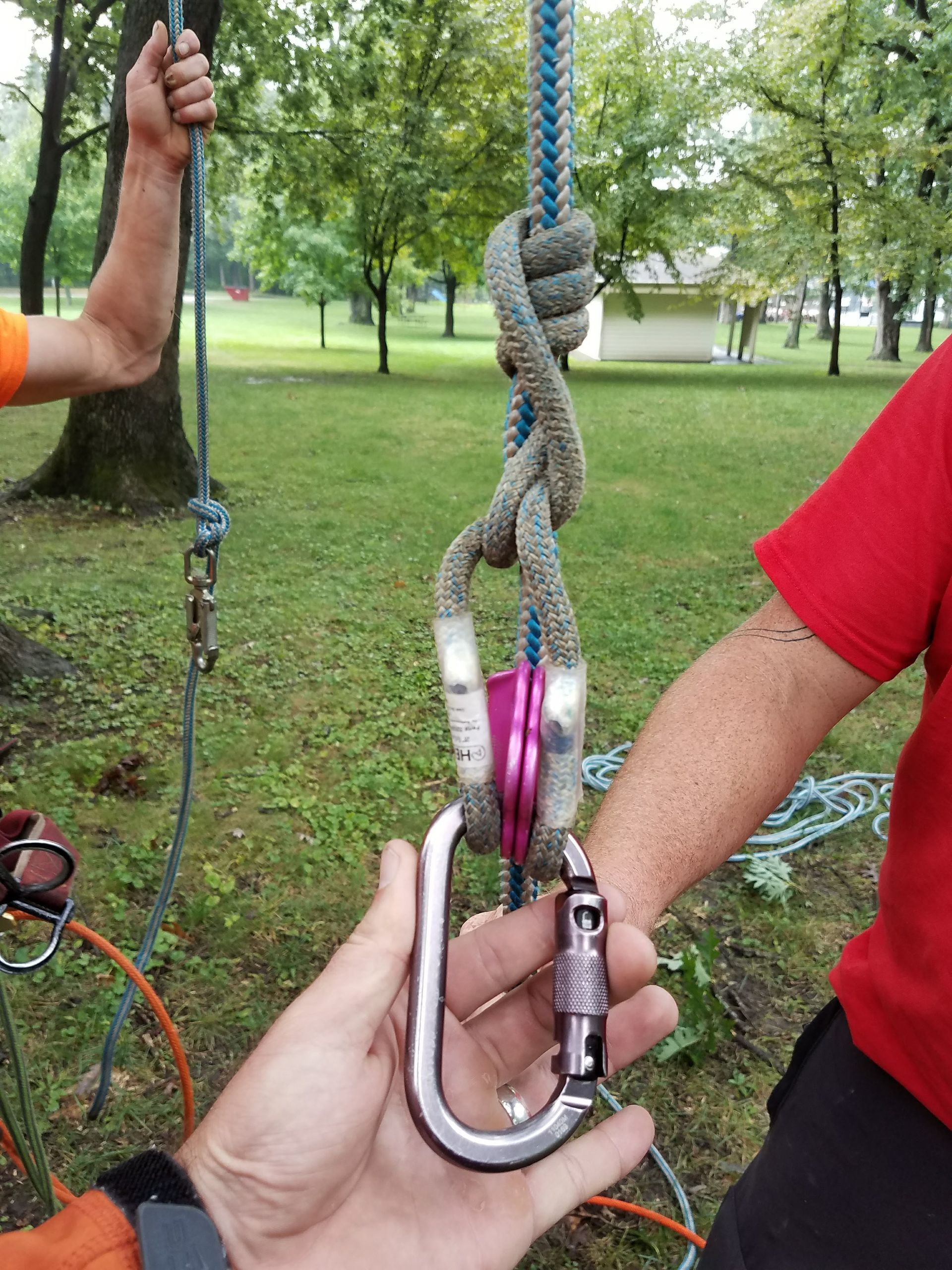 Hands holding carabiner attached to pulley system with rope, outdoors.