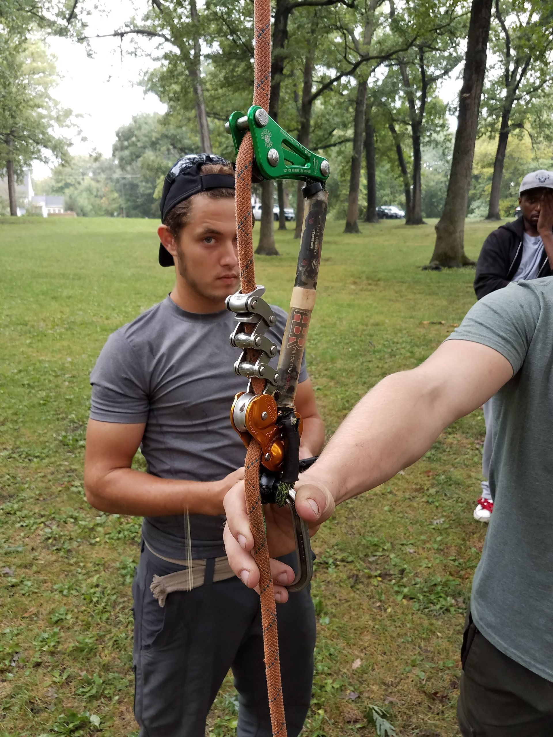 Man holding rope with climbing gear, outdoors. Another person holds rope. Green trees, grassy area.