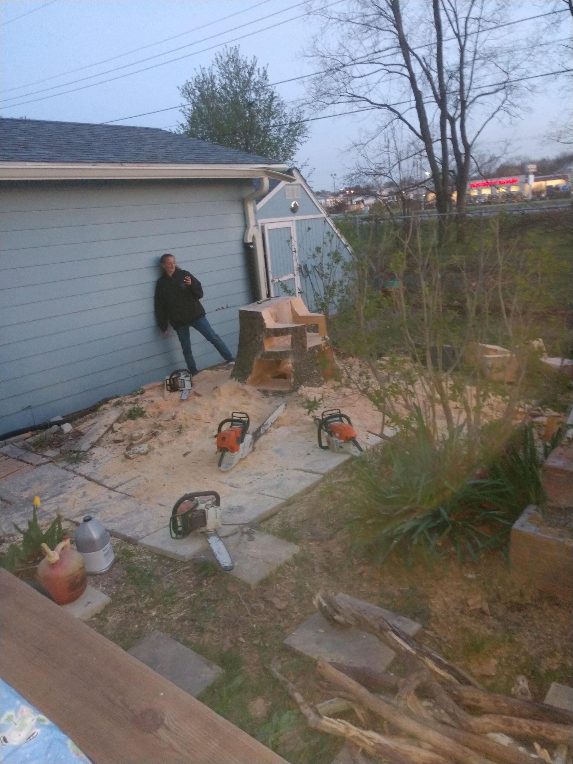 A person leaning on a blue building next to a tree stump. Chainsaws and sawdust are visible in an outdoor setting.