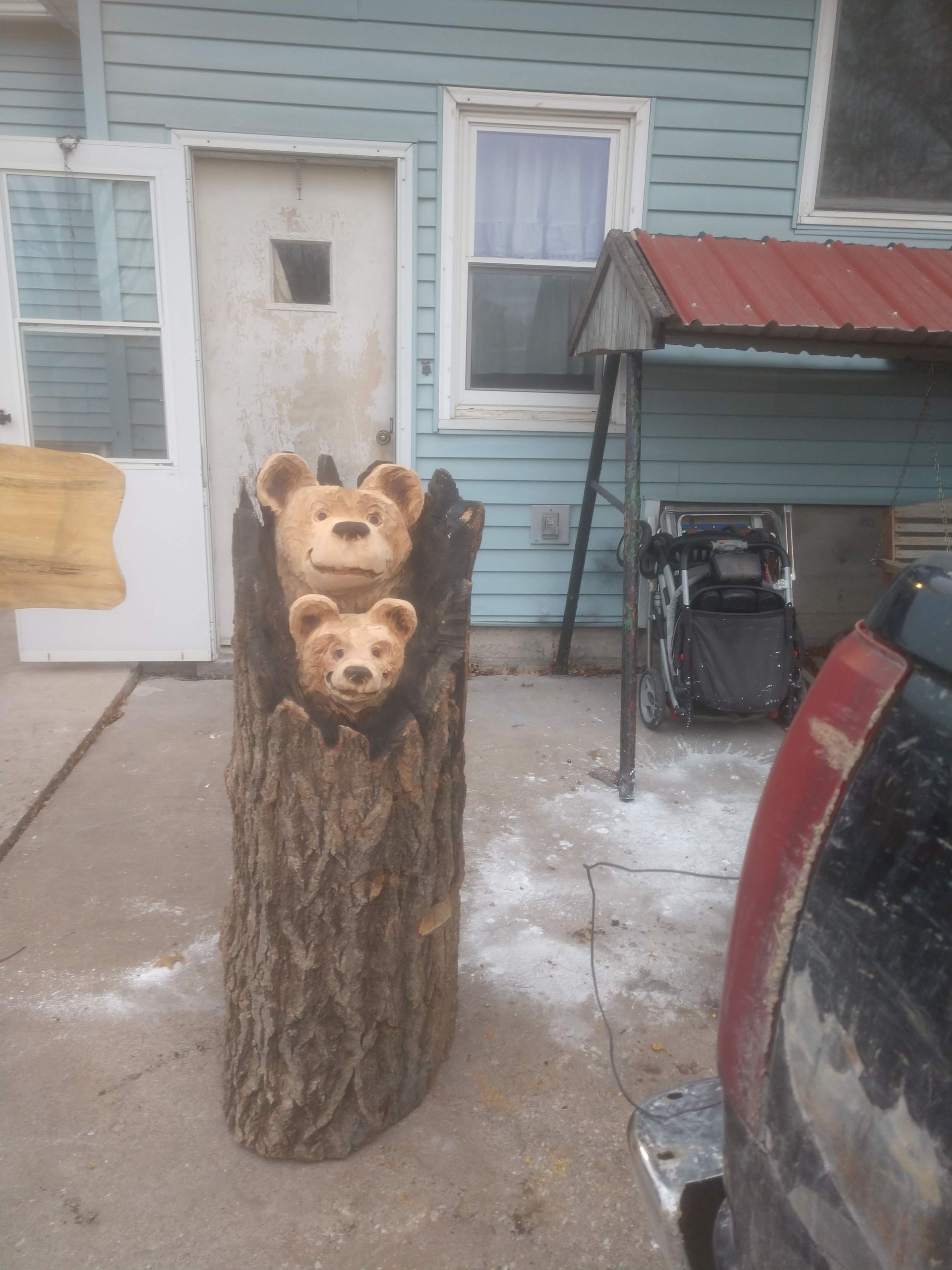 Bear sculpture carved from a tree trunk; two bears peek from the hollow. Outdoors, near a house.