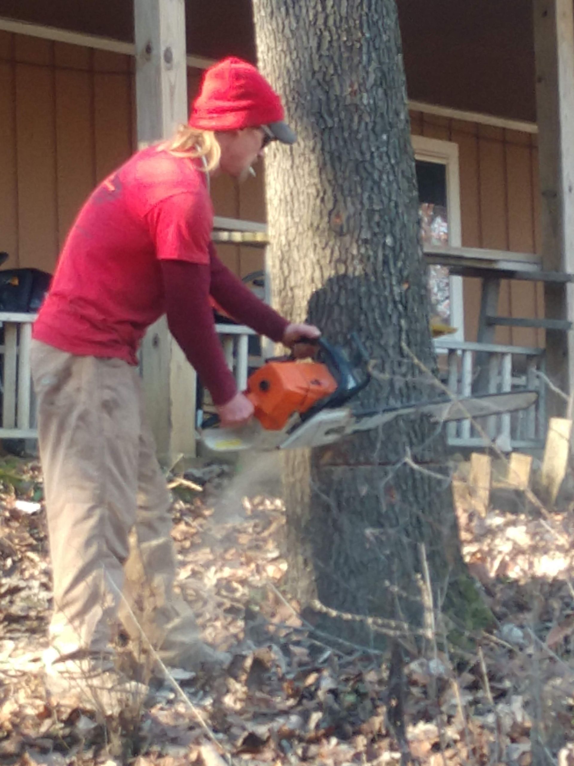Person in red shirt cutting a tree with a chainsaw, outdoors near a porch.