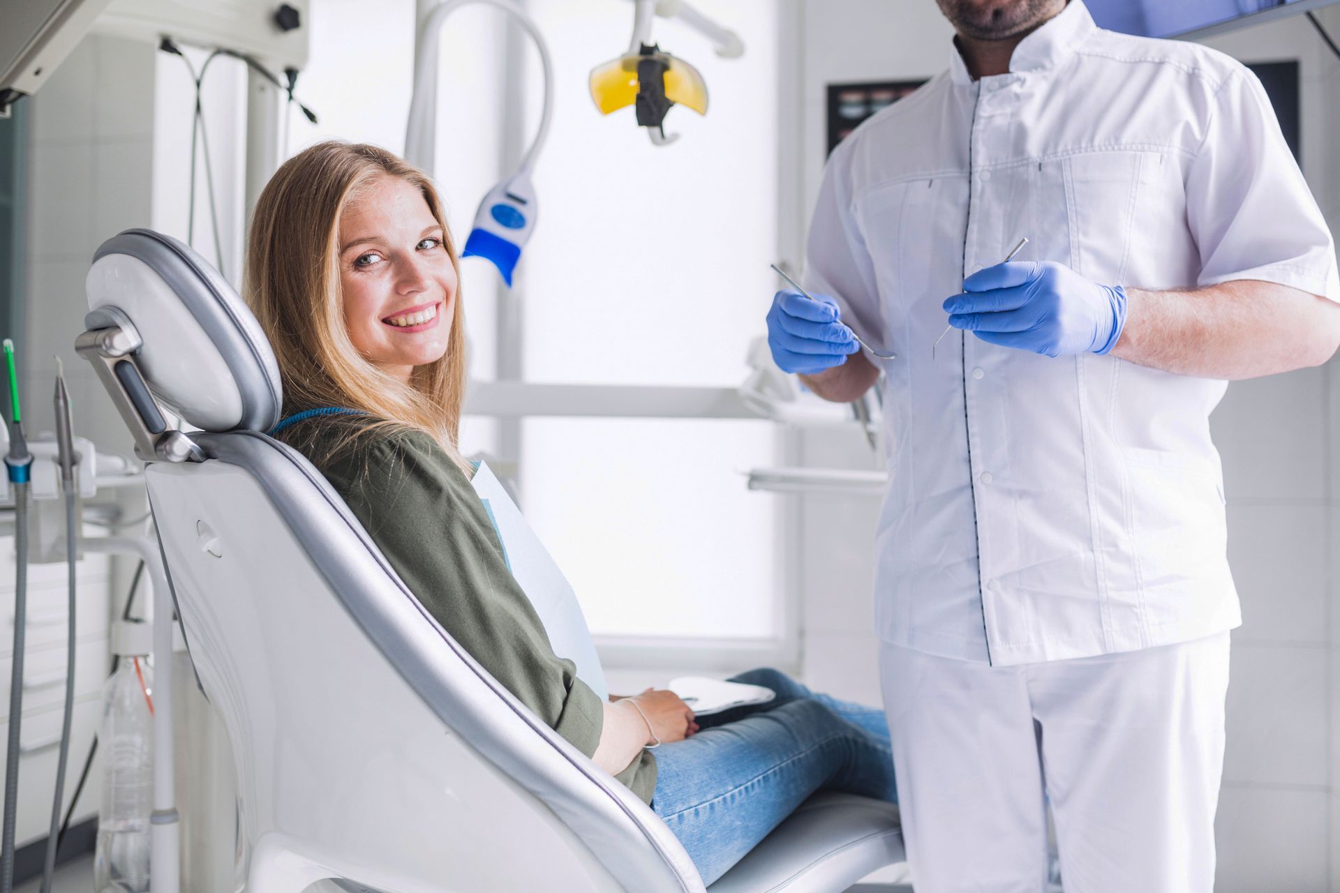 Woman smiles in dental chair; dentist in white uniform and gloves prepares.