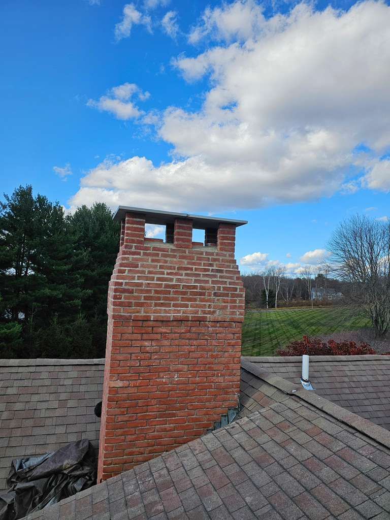Brick chimney on a shingled roof under a blue sky with clouds