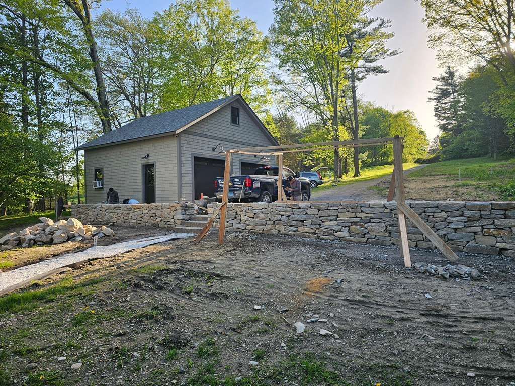 Gray house under construction with gravel yard, stone retaining walls, and trees in the background