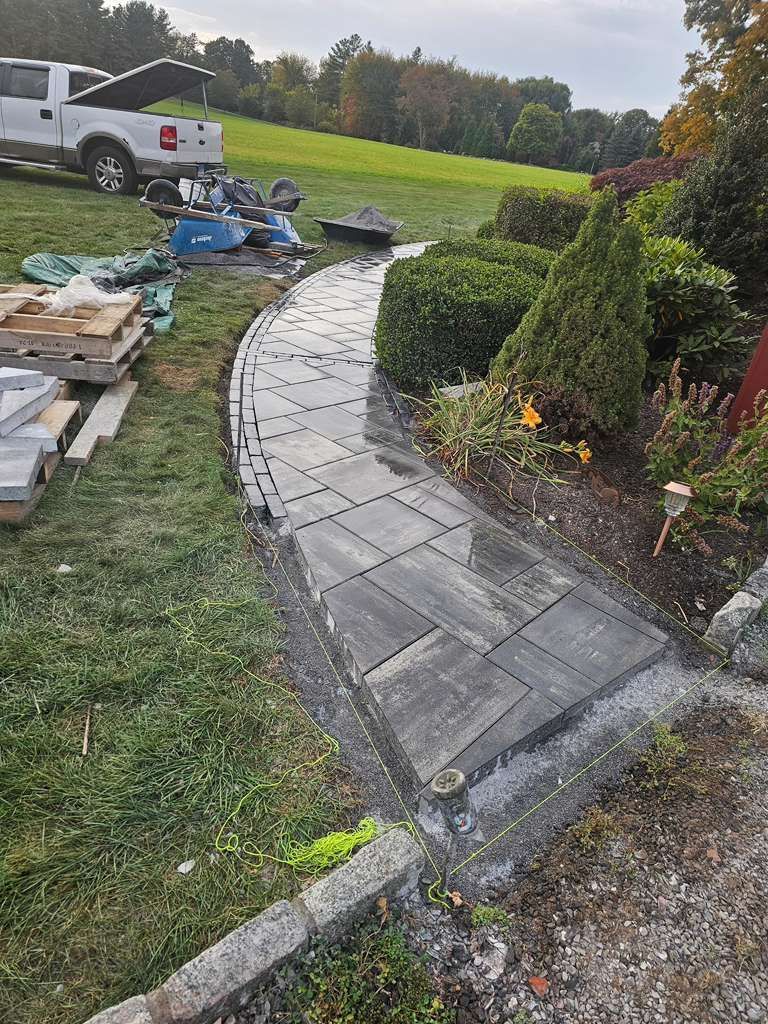 Curved gray paver walkway under construction beside shrubs, with a white pickup and materials on grass