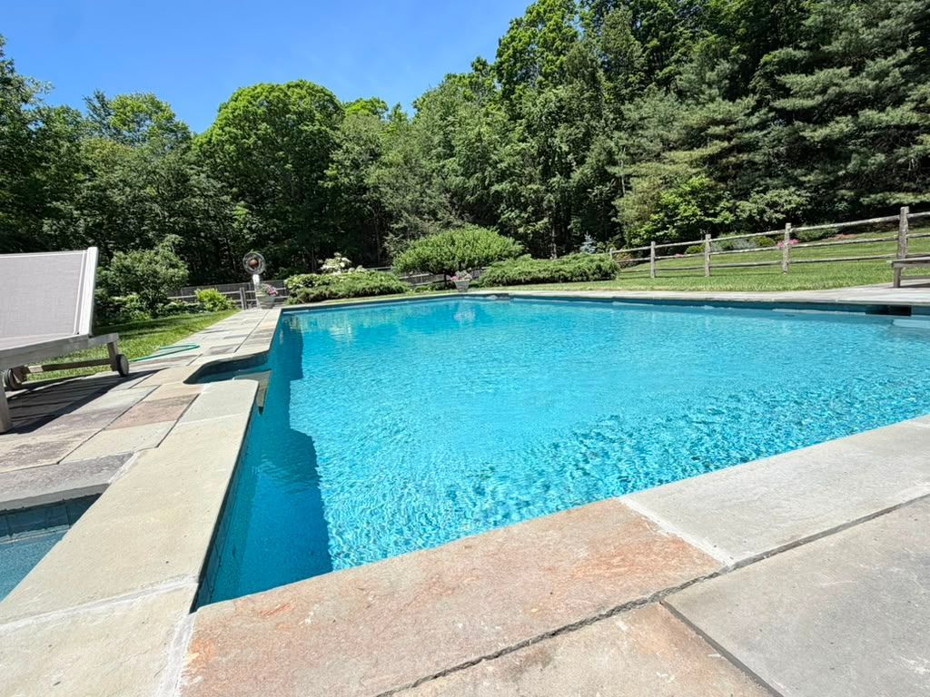 Outdoor swimming pool with turquoise water, stone deck, and trees under a clear blue sky