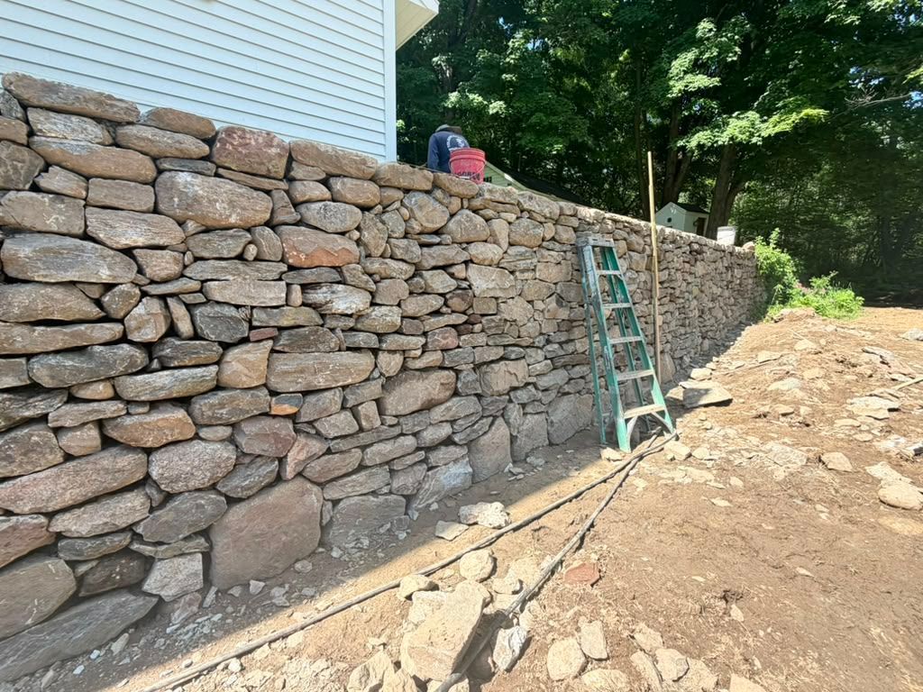 Stone retaining wall beside a house under construction, with a ladder and worker on top.