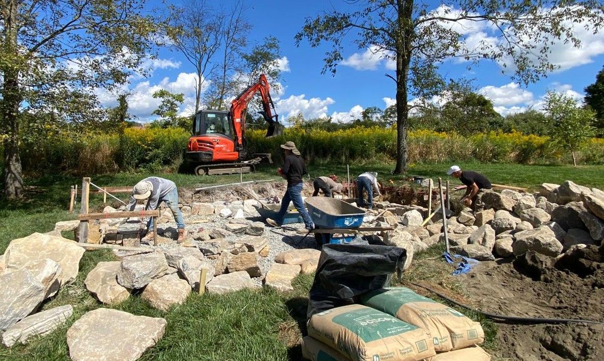Workers building a stone wall beside a red excavator in a sunny outdoor area