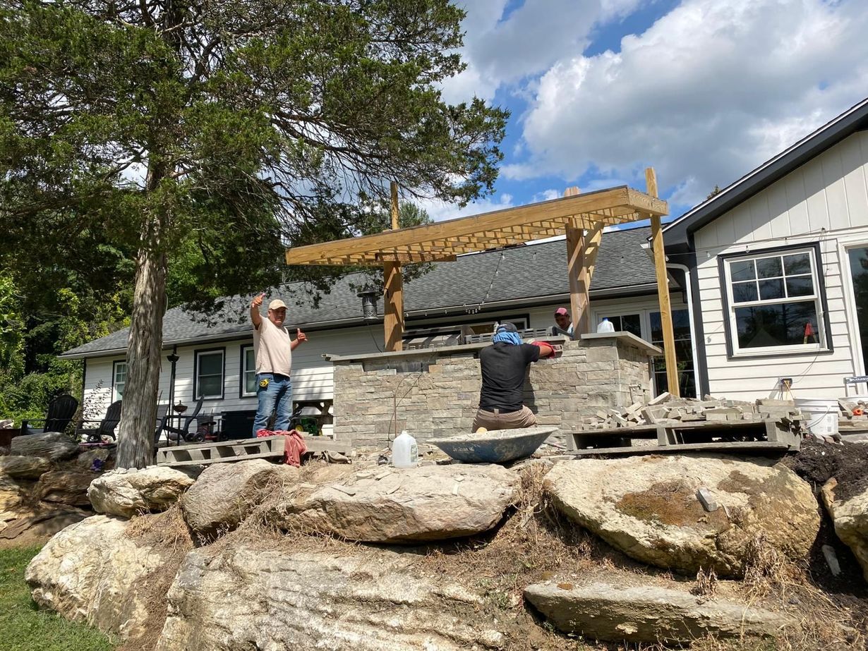 Three people building a pergola beside a stone patio and white house under a blue sky