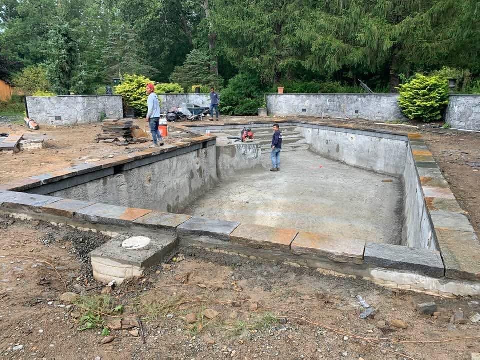 Construction site with an in-ground rectangular pool foundation, workers, and trees in the background