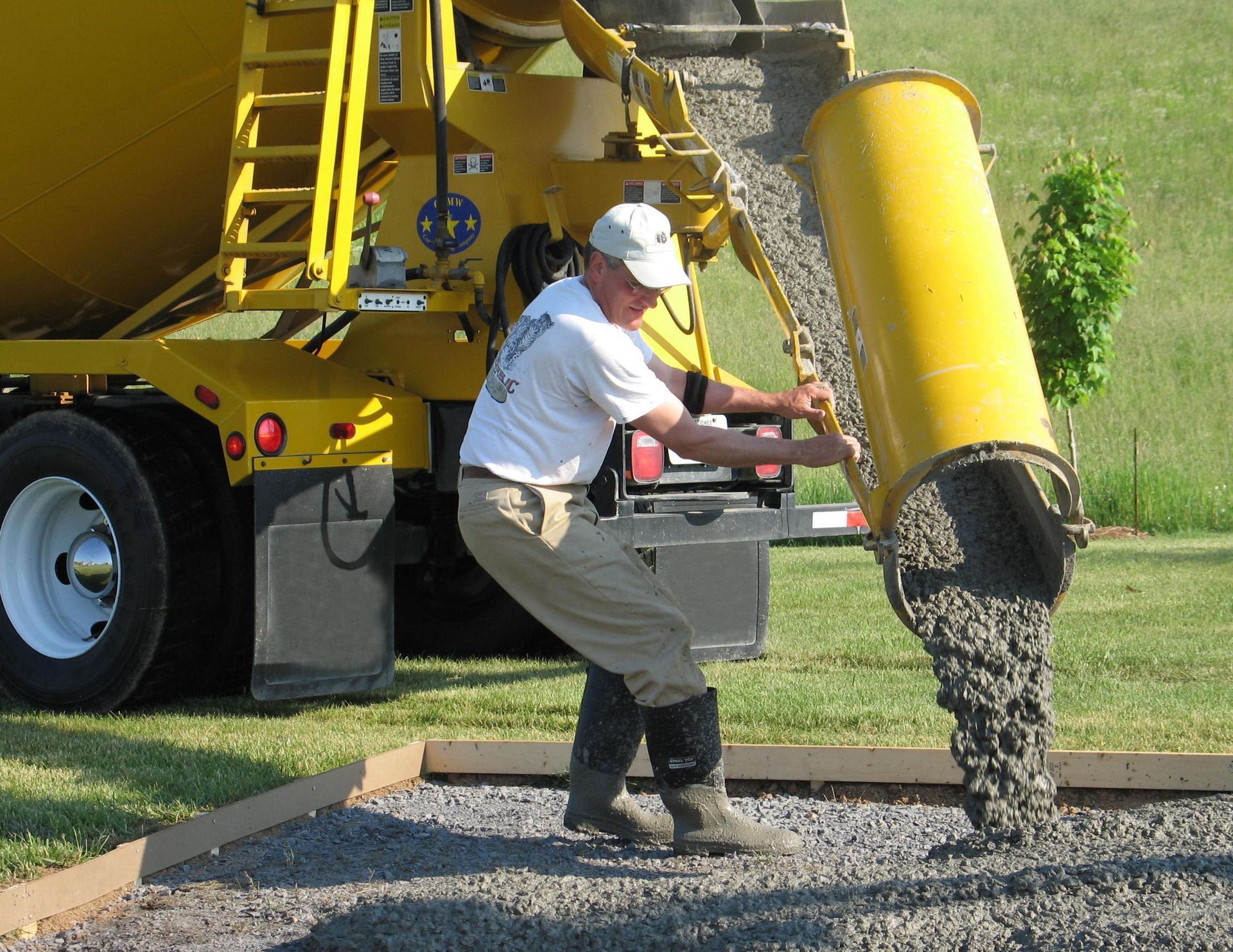 Man pours wet concrete from a yellow truck's chute into a framed area outdoors.