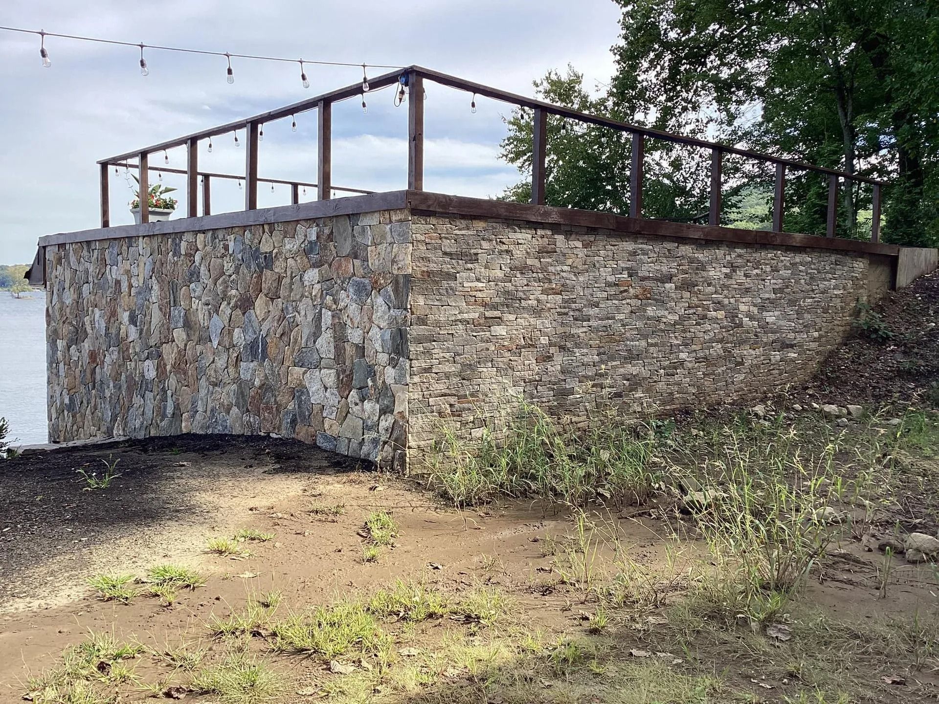 Stone-walled platform with metal railings overlooking a body of water.  Dirt and some vegetation in the foreground.