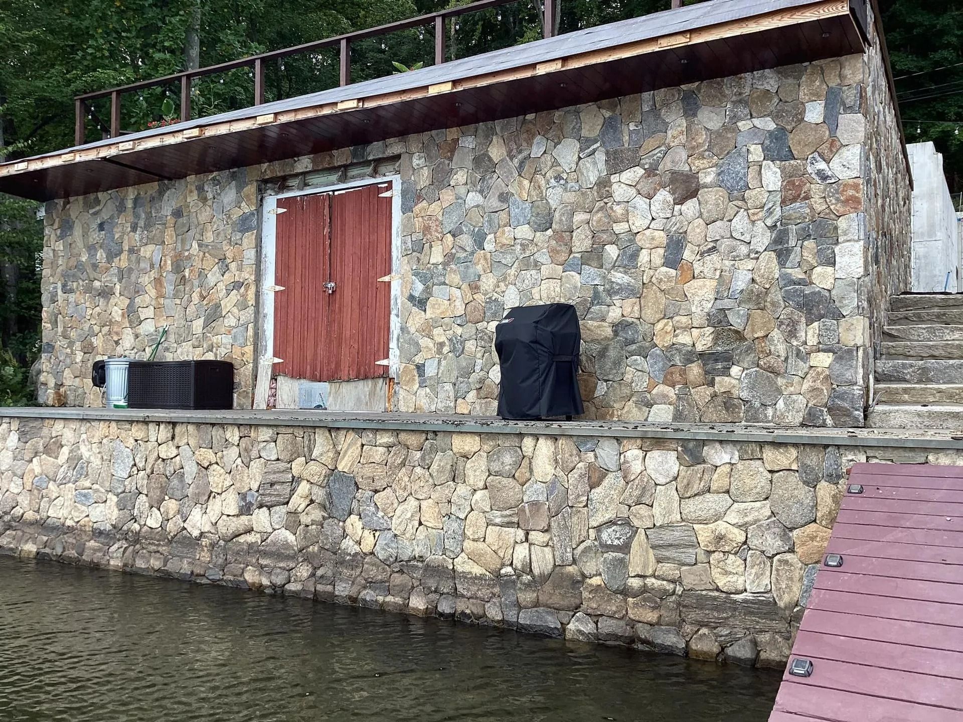 Stone building with red door by water, steps lead to the right. A dark object is covered and placed in front of the door.