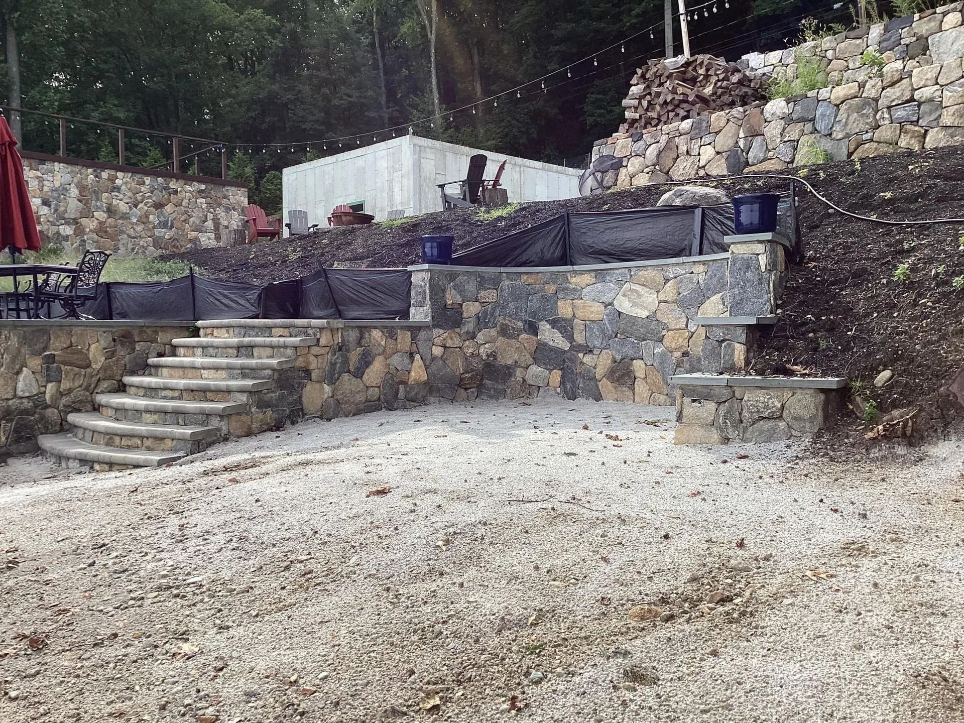 Stone patio with steps and retaining walls, gravel ground. Dark seating area, lush green background.