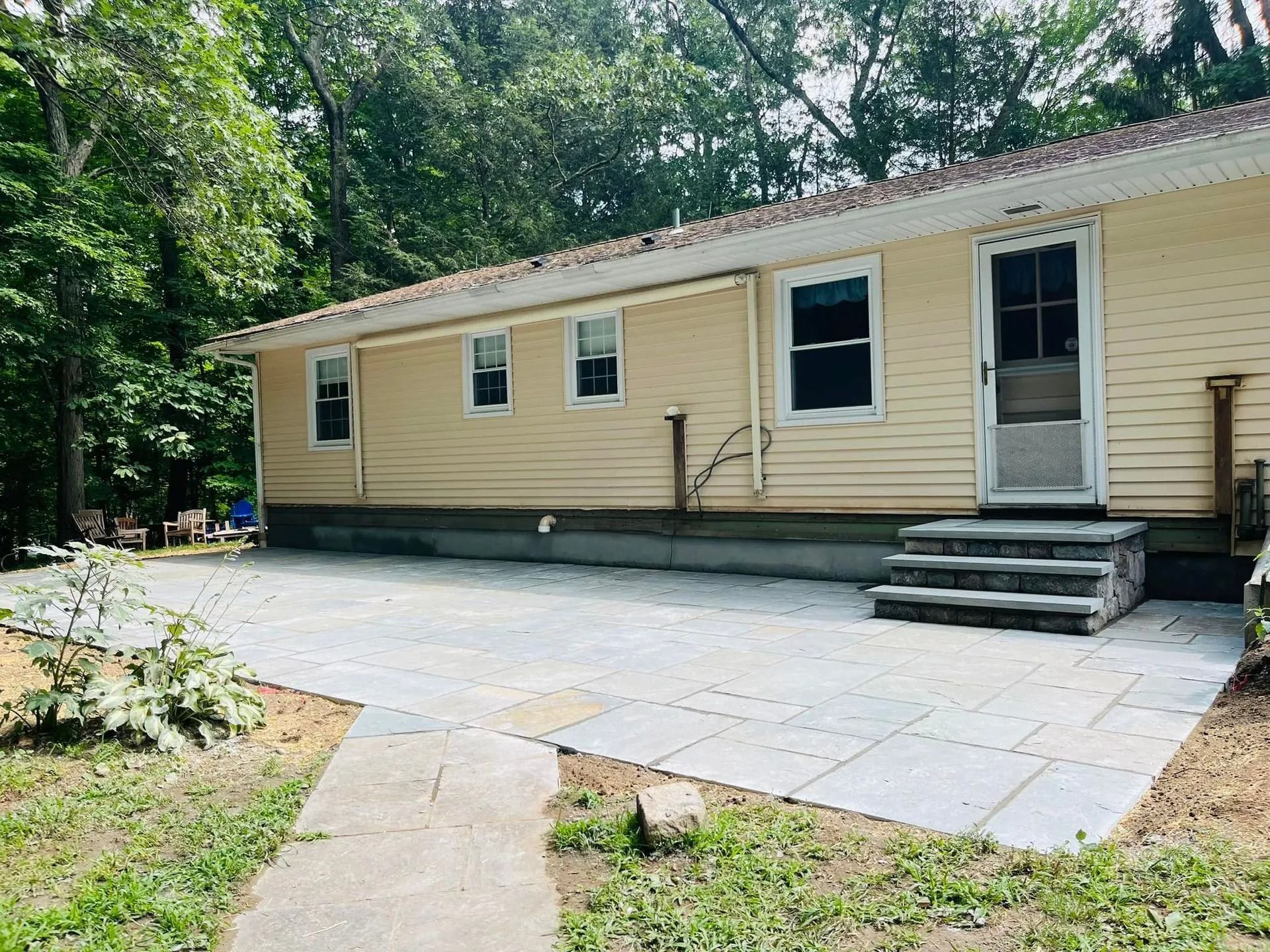 Beige house with a patio, steps, and door. Gray paving, green lawn, and trees surround it.