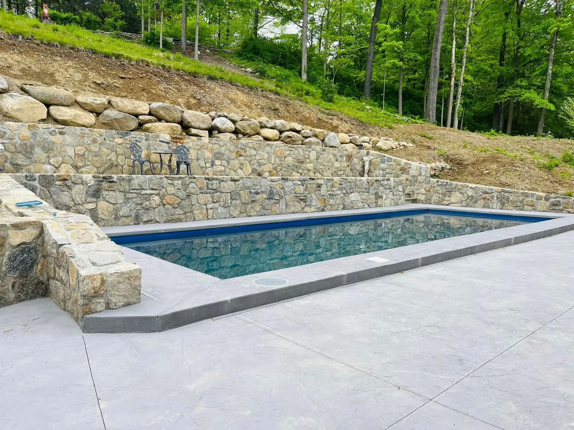 Stone-walled pool with a gray concrete deck, set against a hill with trees.