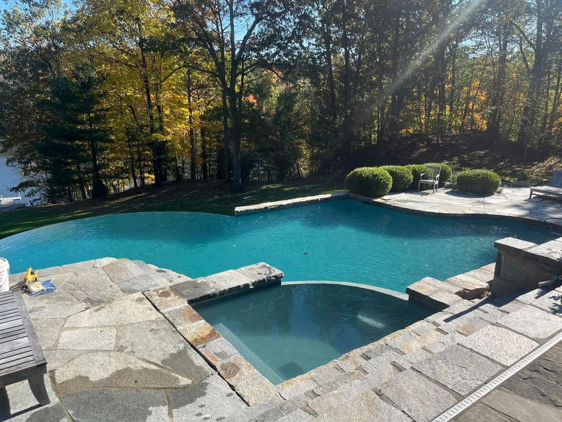 Swimming pool with spa, stone patio, and trees in background.