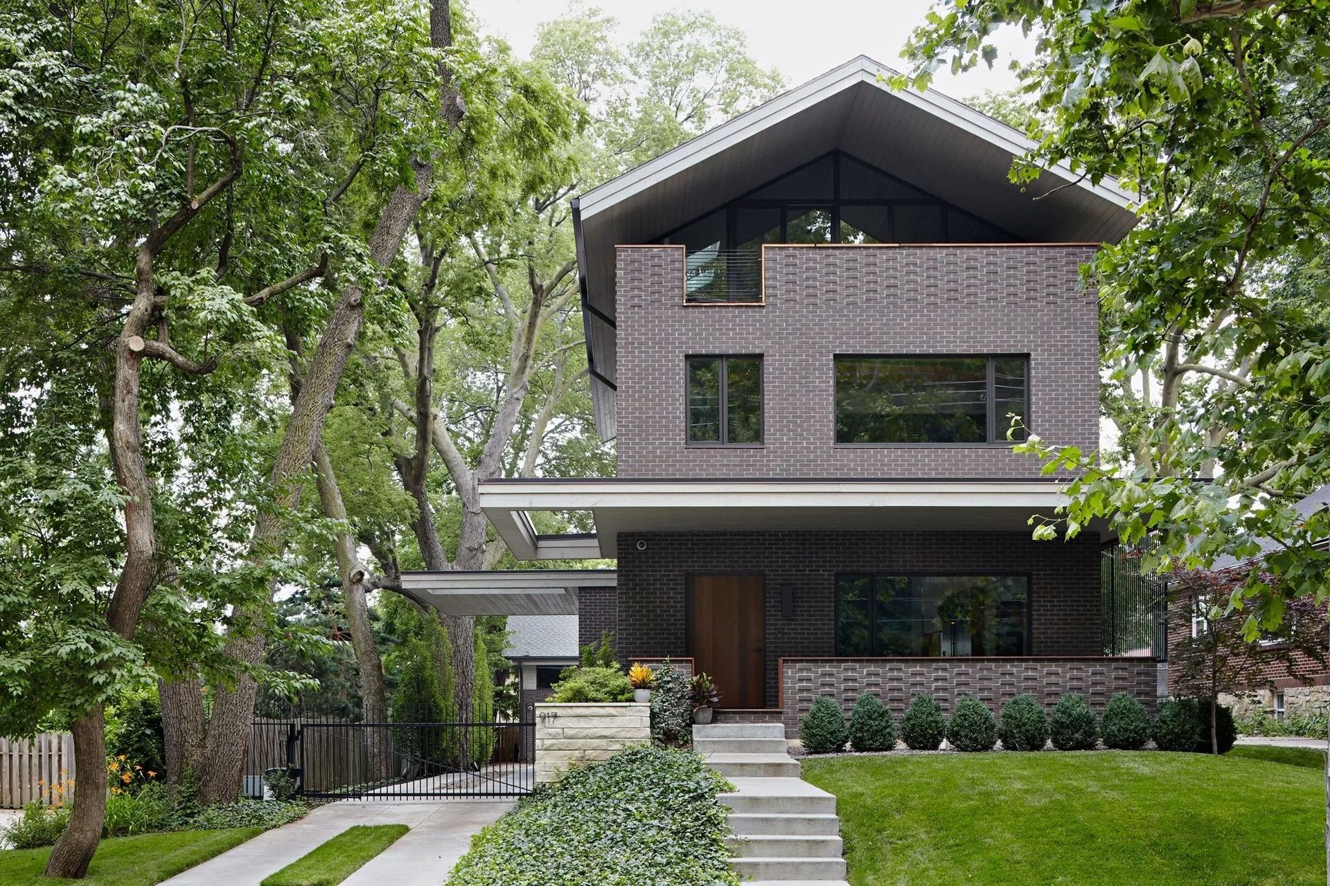 Modern two-story brick house with a gabled roof and a balcony, set among lush green trees.