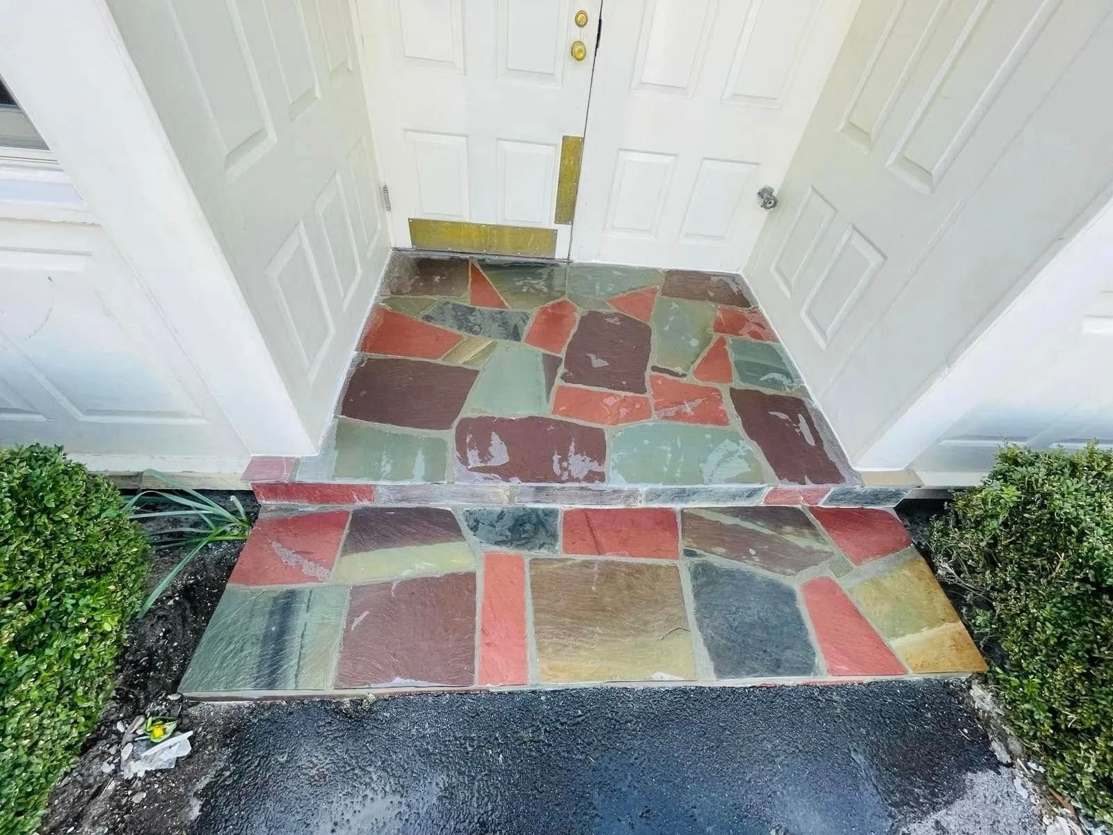 Stone-tiled porch entrance with colorful, irregularly shaped tiles; white door and trim; small bushes on either side.