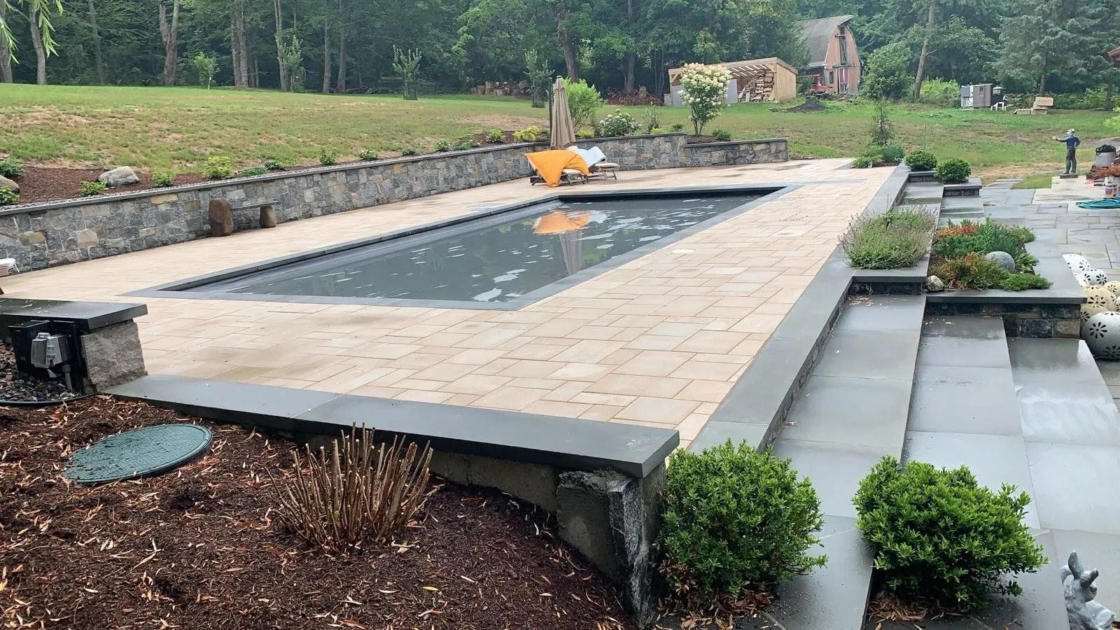 A rectangular in-ground pool surrounded by beige pavers and stone retaining walls. Green foliage and a grassy lawn are in the background.