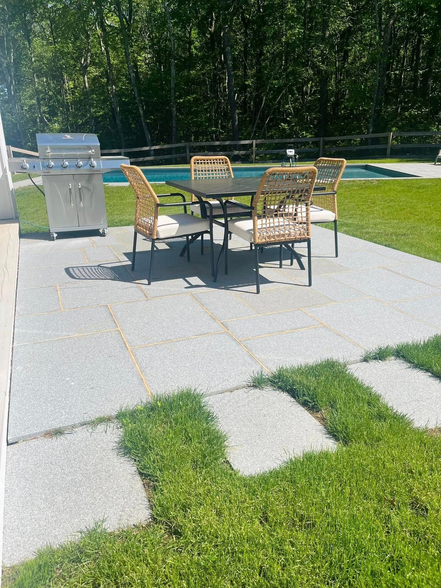 Patio with dining set, grill, and swimming pool in the background. Green grass and trees surround the area.