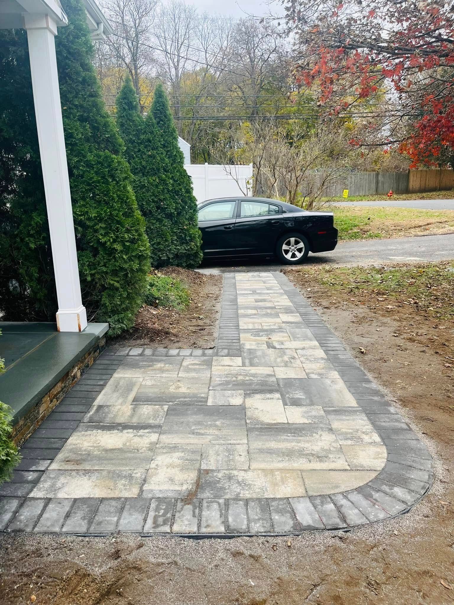 Paver walkway leading to a porch. Black car parked in the background, green bushes and colorful trees.