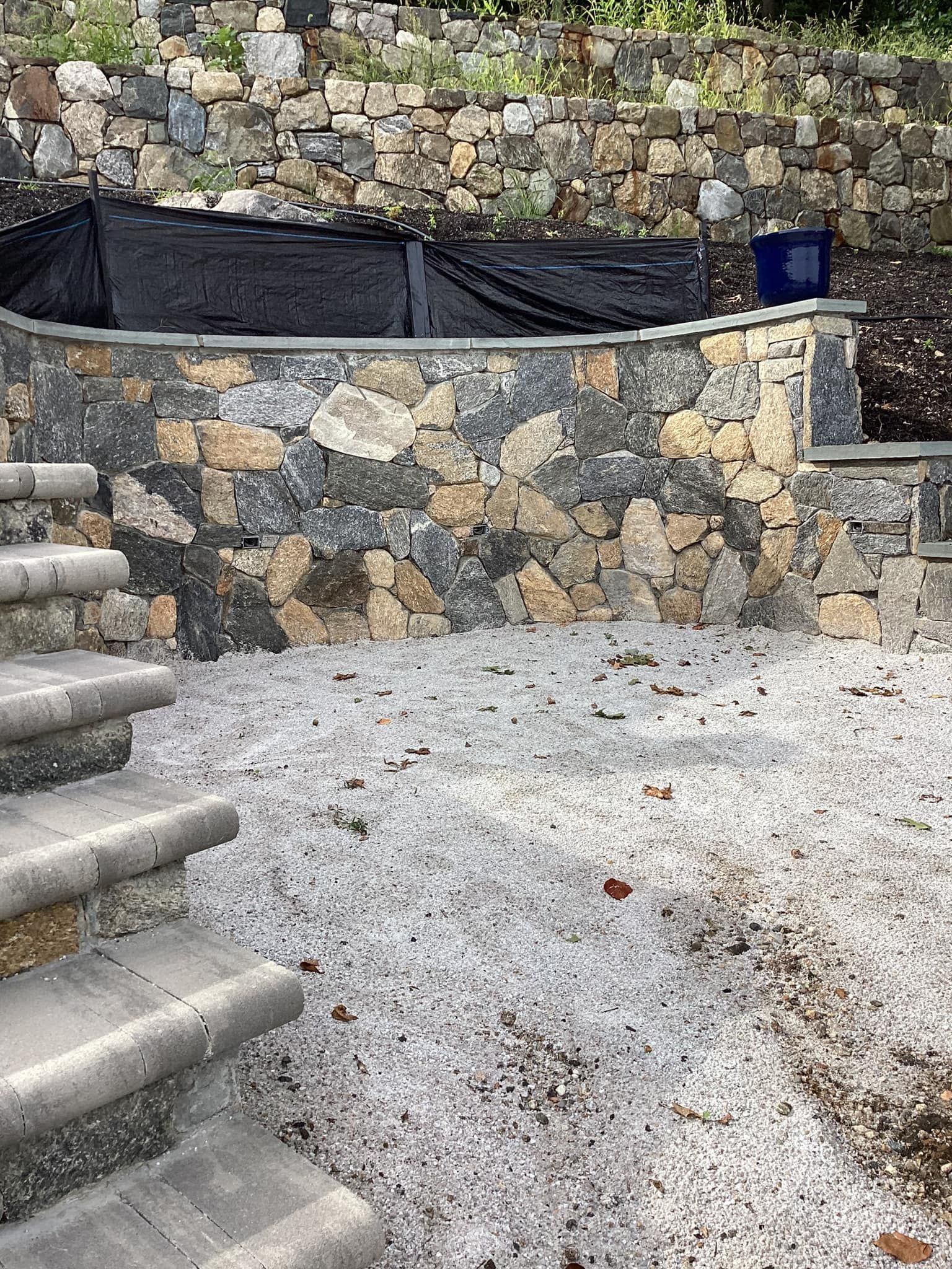 Stone wall and steps leading to a gravel-covered courtyard with a retaining wall and dark fabric covering.