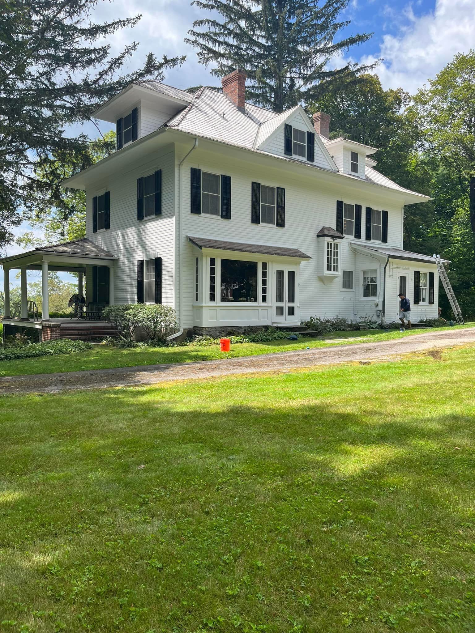 White two-story house with black shutters, on a grassy lawn. Porch on left. Green trees in the background.
