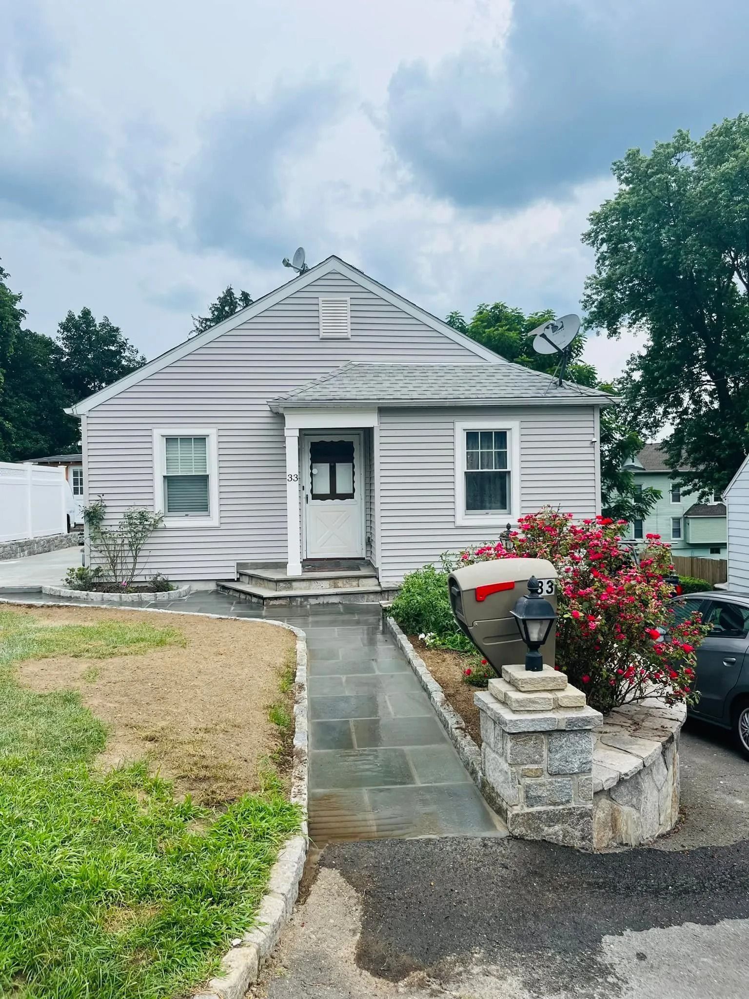 Gray house with a walkway, small front yard, and bushes, under a cloudy sky.