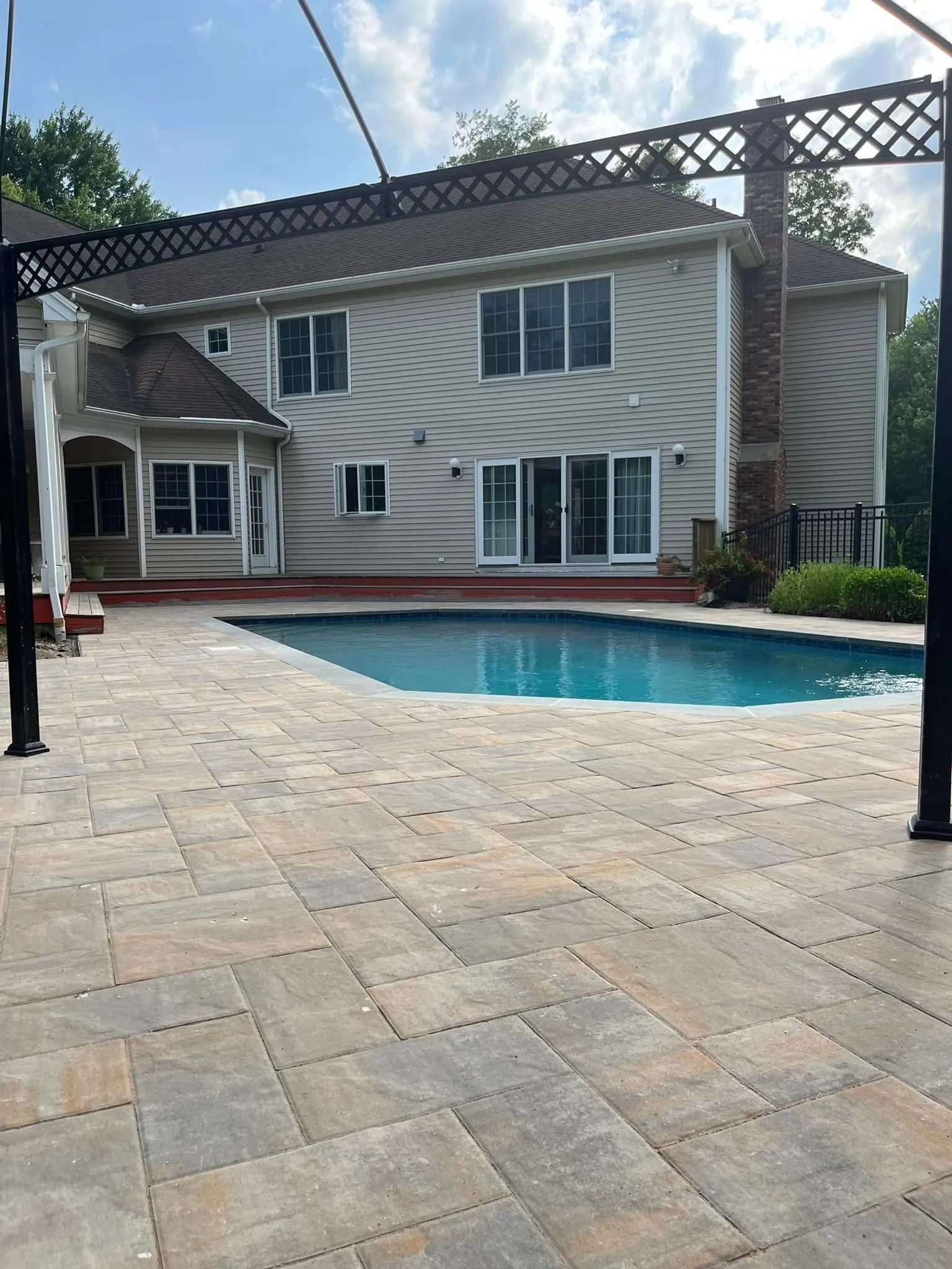 Backyard with pool and house. Beige pavers surround the pool. House has light siding, a red deck, and a chimney.