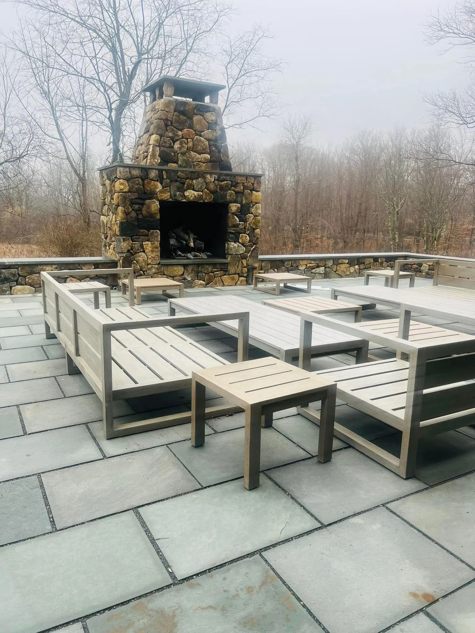 Outdoor patio with stone fireplace and light-colored wooden seating on gray pavers.