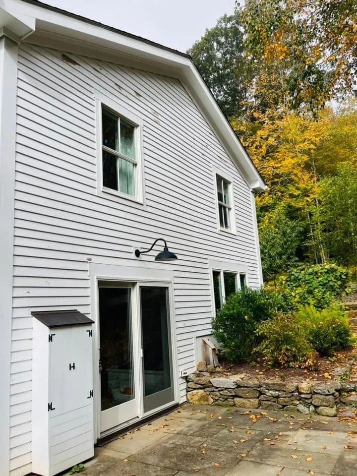 White clapboard house with black awning light and glass doors, surrounded by trees and stone wall.