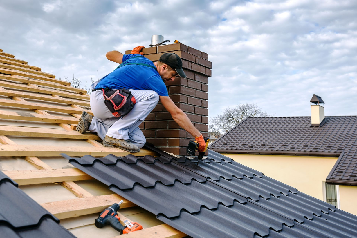 Roofer installing dark metal roofing near a brick chimney on a cloudy day.