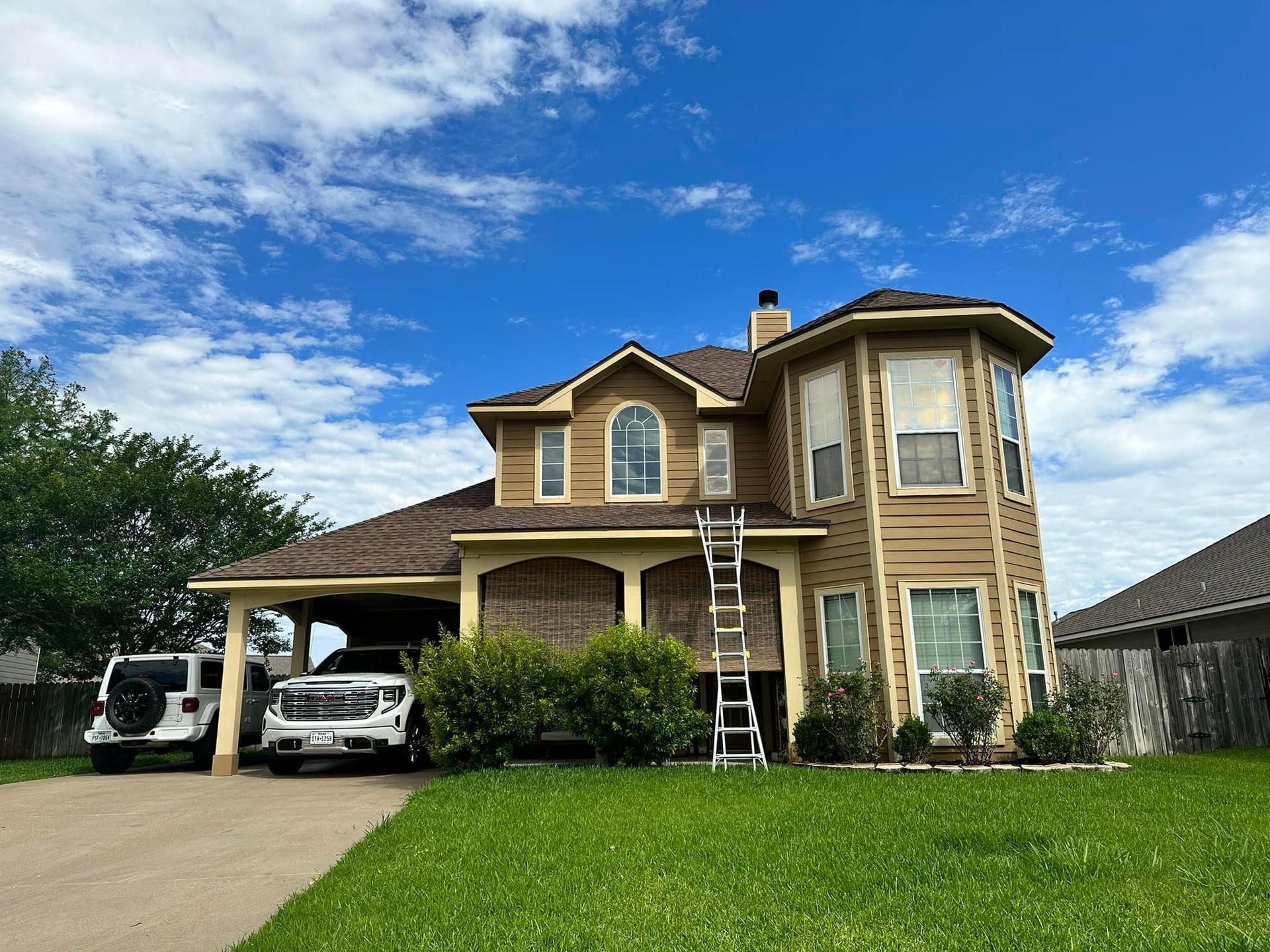 Two-story house with tan siding, brown roof, and cars parked in the driveway. A ladder leans against the building.