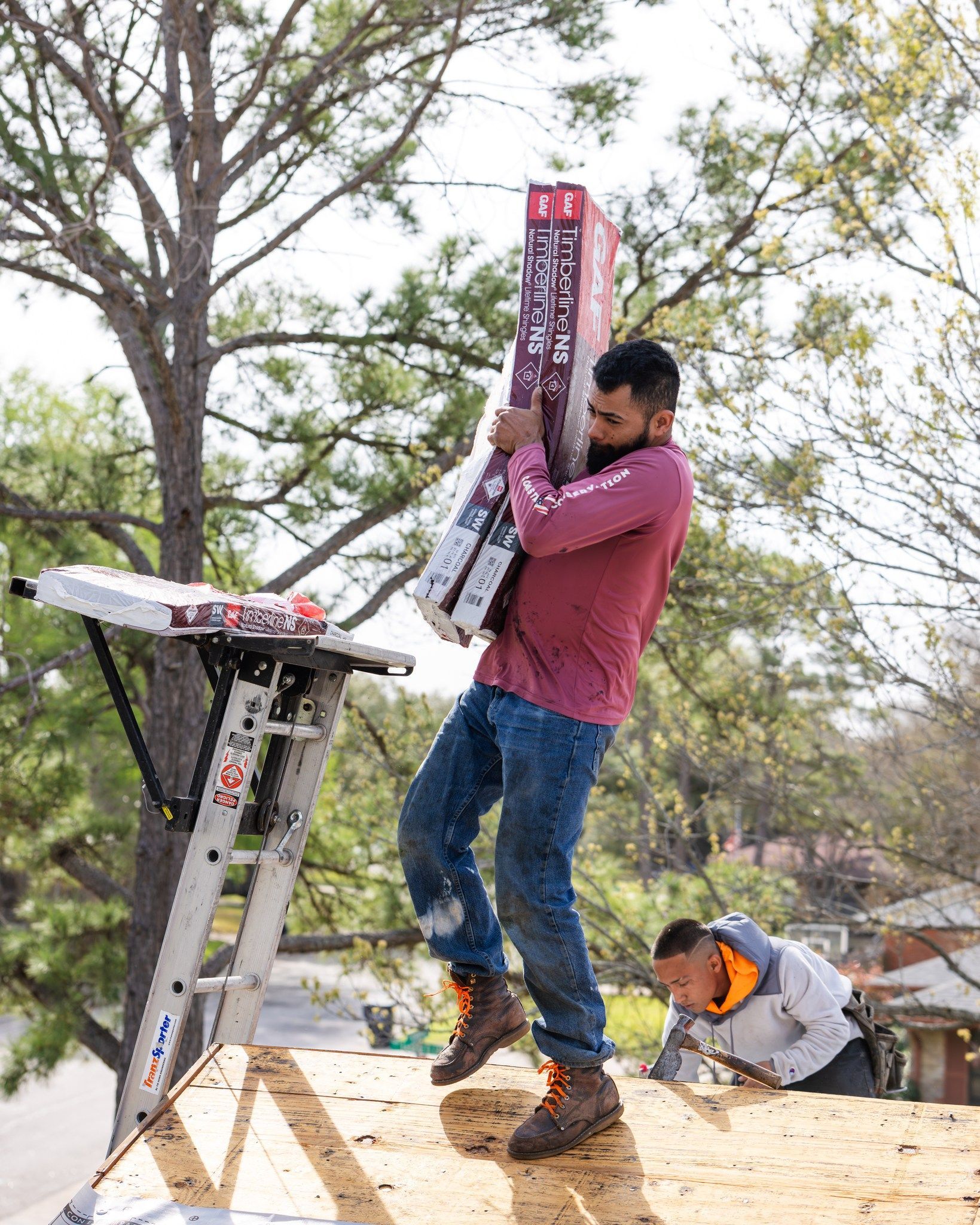 Construction worker carrying roofing materials up a ladder. Another worker on roof. Outdoors.