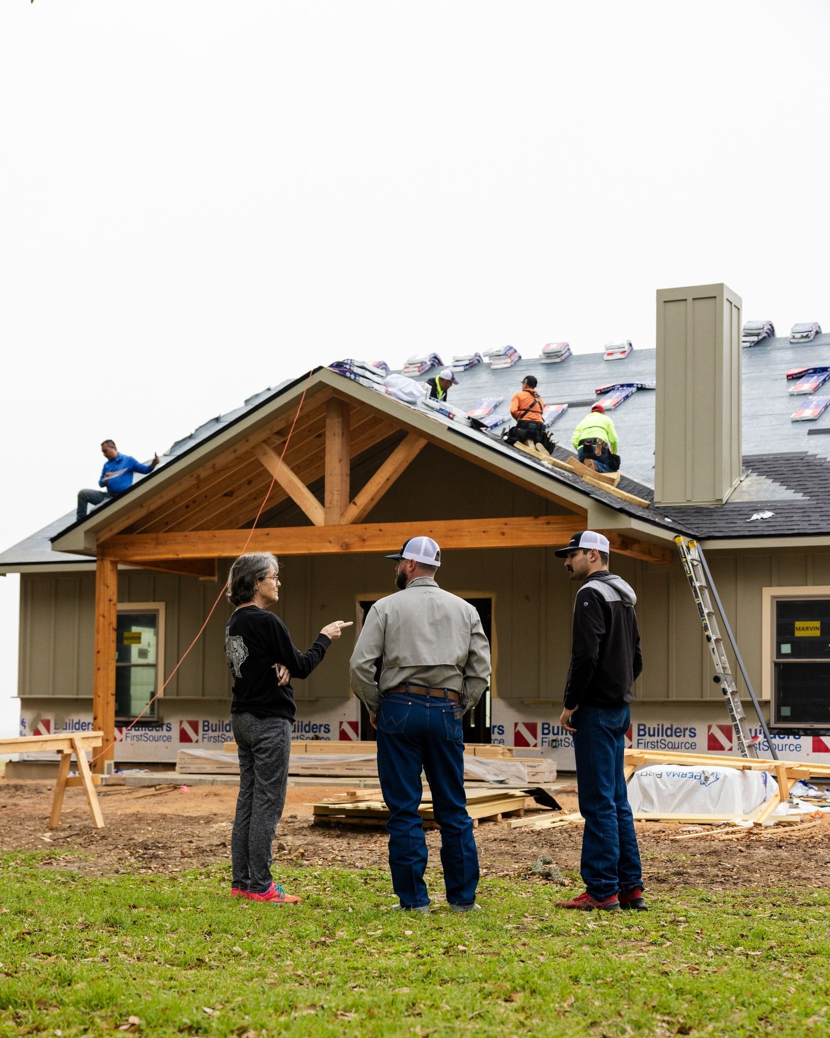 Construction workers on a roof, talking to three people in front of a house under construction; cloudy day.
