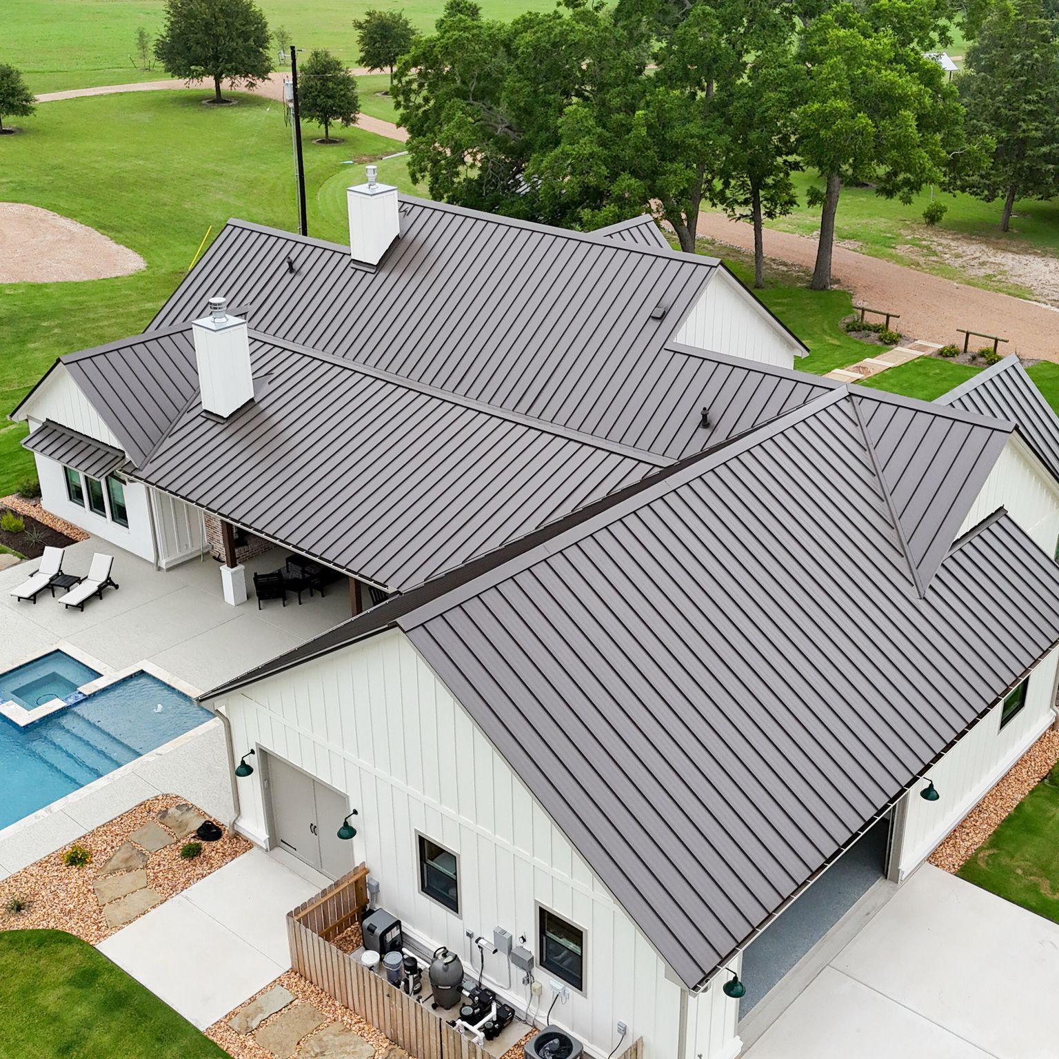 Aerial view of a white house with a gray metal roof, a pool, and a garage in a green, grassy setting.