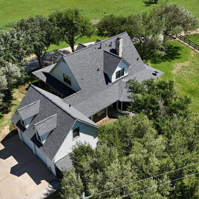 Aerial view of a gray-roofed house with dormers, surrounded by trees and green grass.
