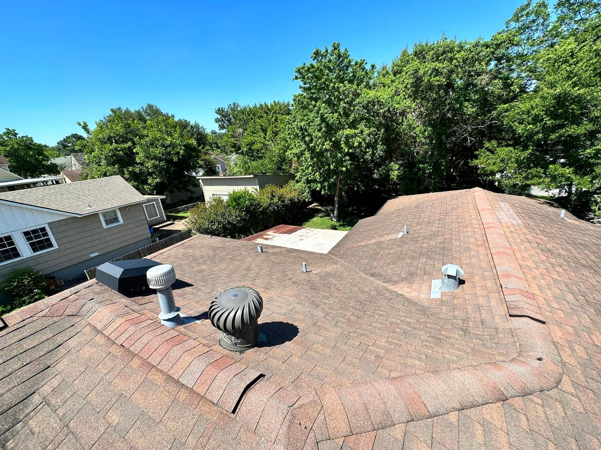 Overhead view of a house roof with a spinning ventilator. Trees and blue sky are in the background.