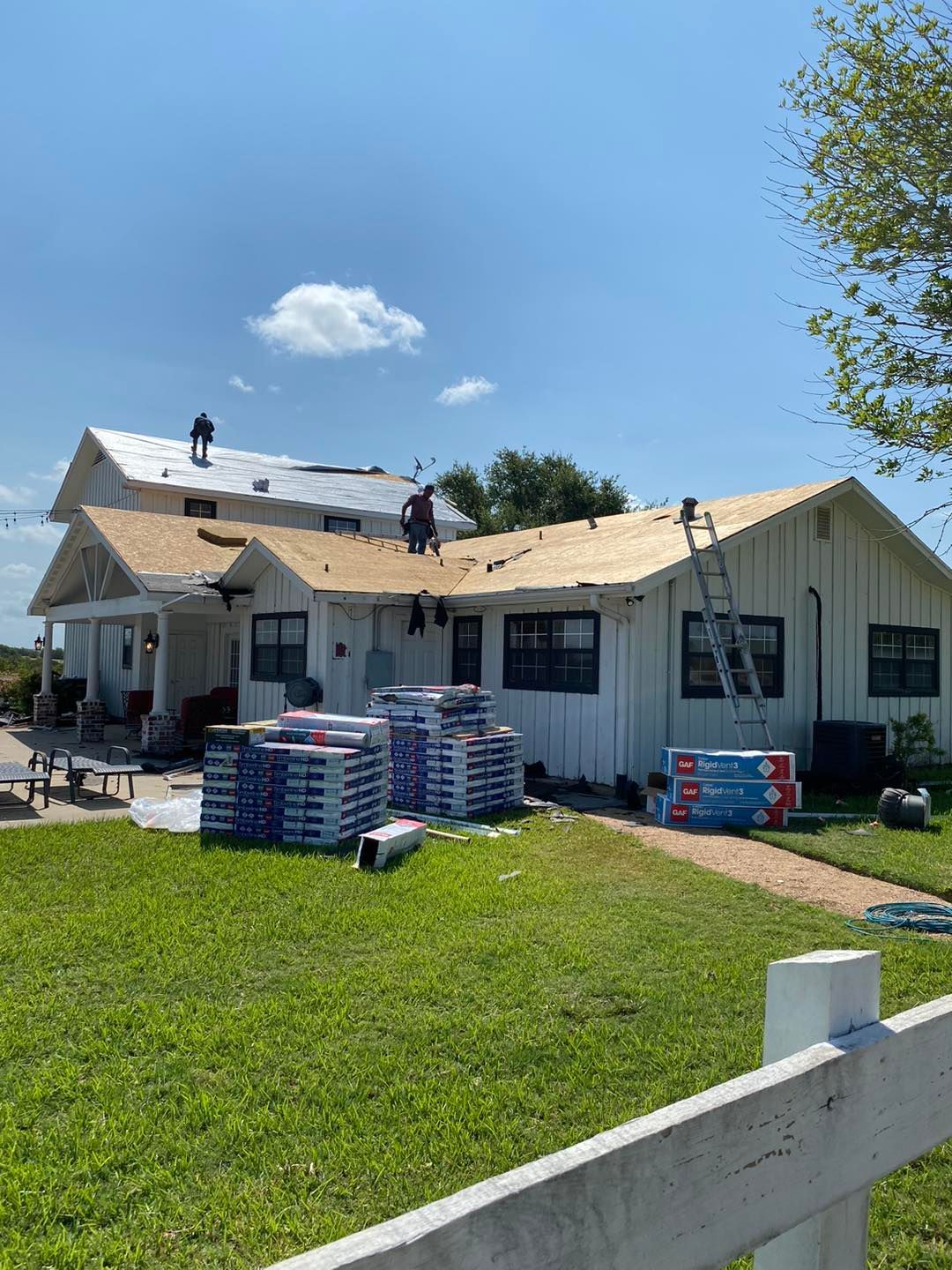Workers replacing roof shingles on a white house. Shingles and ladder on the ground. Sunny day.