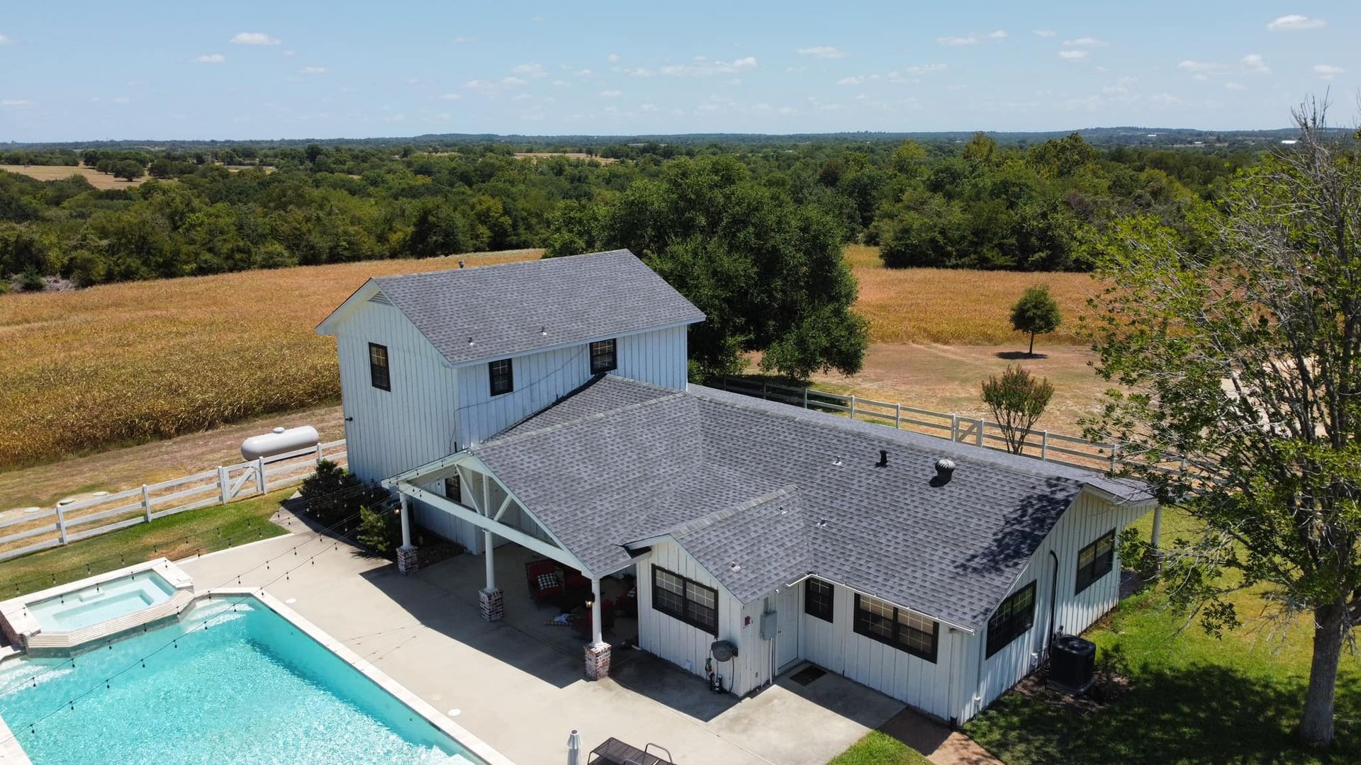 Aerial view of a white farmhouse with a pool, surrounded by fields and trees under a blue sky.