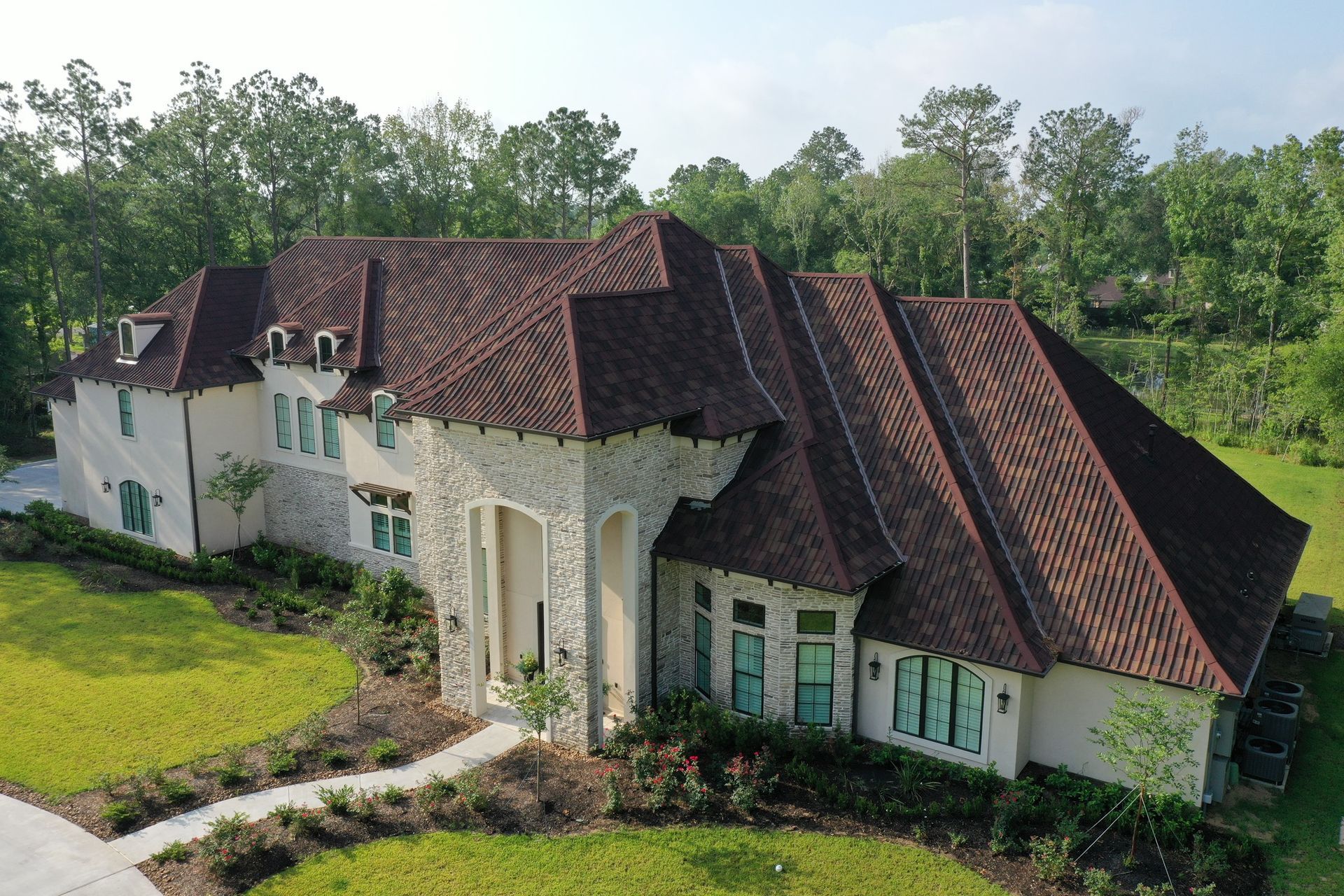 Large beige and stone house with a red-brown tile roof, surrounded by green grass and trees.