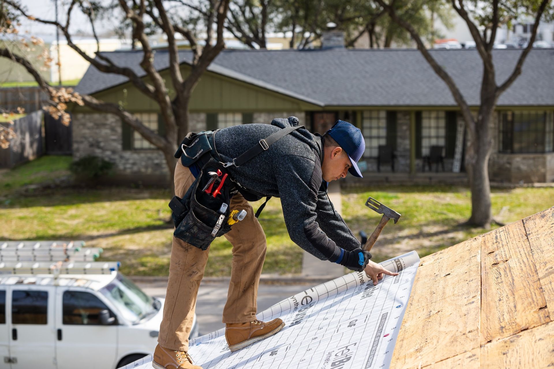 Roofer in blue cap hammering roofing material on a residential roof; white van and house in background.
