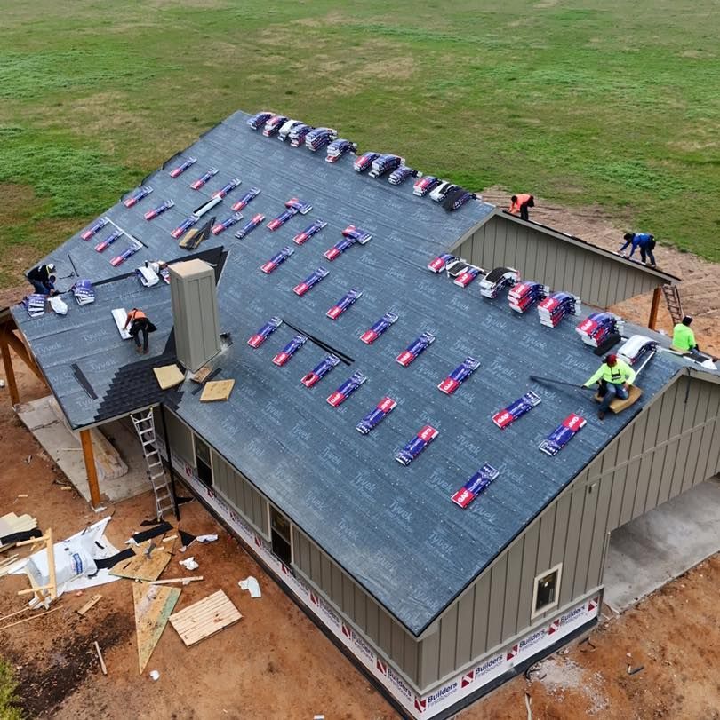 Roofers installing shingles on a house with a gray roof and tan siding.